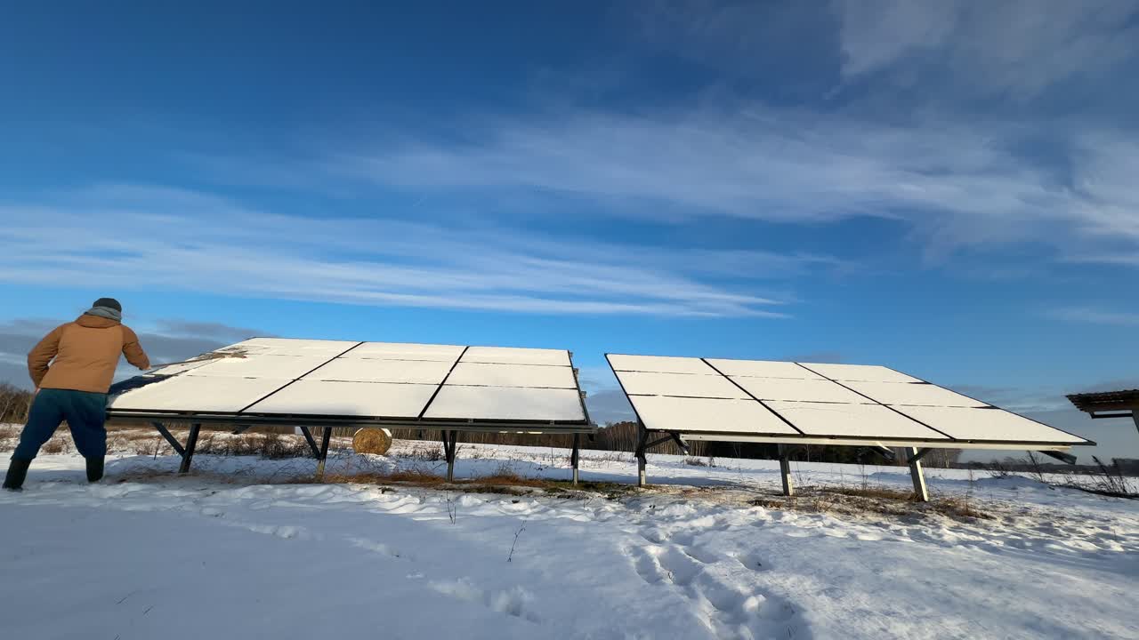 Slow motion begins with panels fully buried in snow. A person starts brushing the first strip clean using a long-handled broom, revealing dark cells under bright frosty winter light