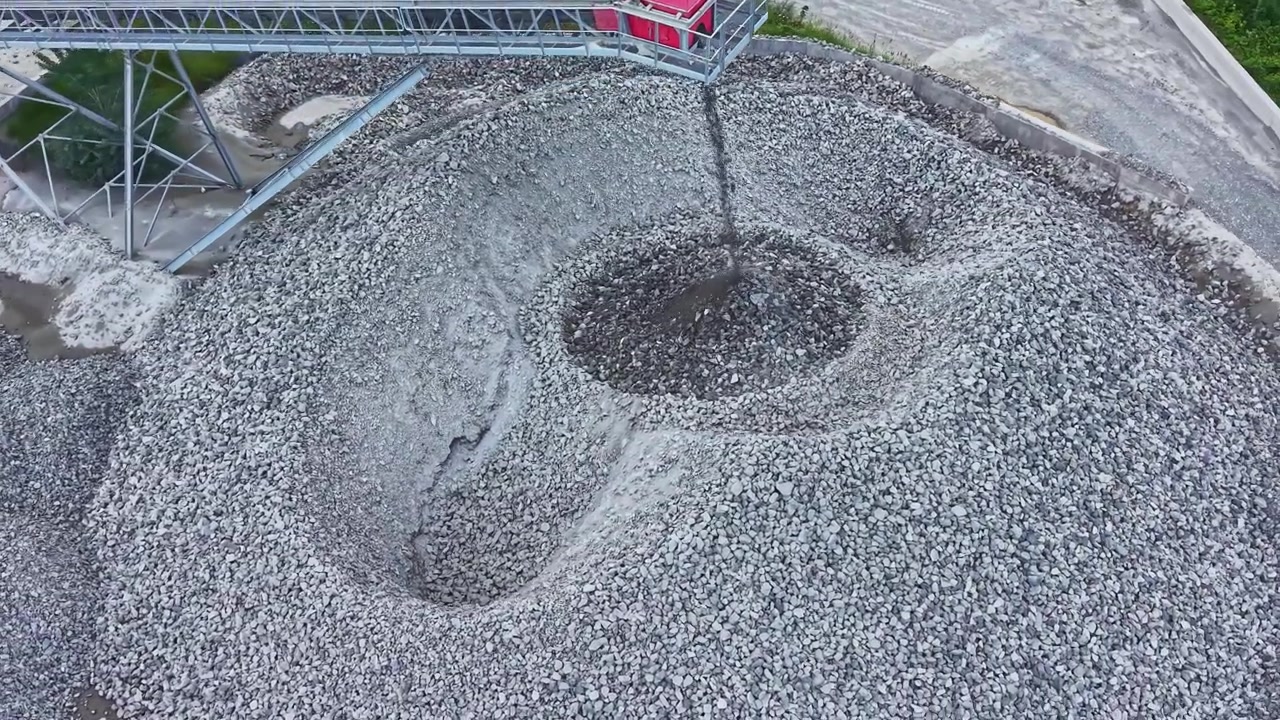 Overhead shot of crushed stone. Drone captures spiral pattern of gravel mound. Aerial perspective reveals spiral gravel formation amid machinery and green embankment