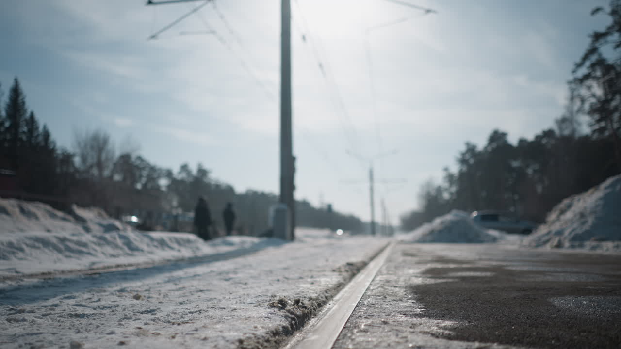 long shot close view of rail cutting through snow along winter tram route, low perspective beside platform shows icy texture and sun glare, distant figures walking, quiet urban stop mood