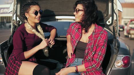 Dos-Jóvenes-Mujeres-Atractivas-Con-Elegantes-Gafas-De-Sol-Sentadas-En-El-Baúl-Del-Camión-En-El-Estacionamiento-Junto-Al-Centro-Comercial-Y-Hablando-Durante-El-Día-Soleado