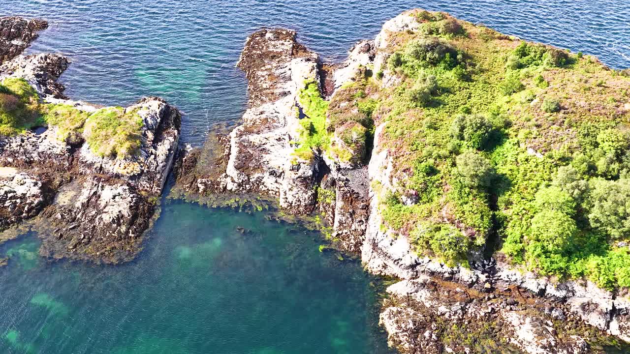 Drone glides over rocky island with lighthouse, clear coastal waters, and lush greenery in sunlight