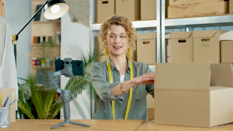 caucasian young woman recording video for fashion blog on smartphone camera sitting in own clothing shop studio while unpacking clothes boxes