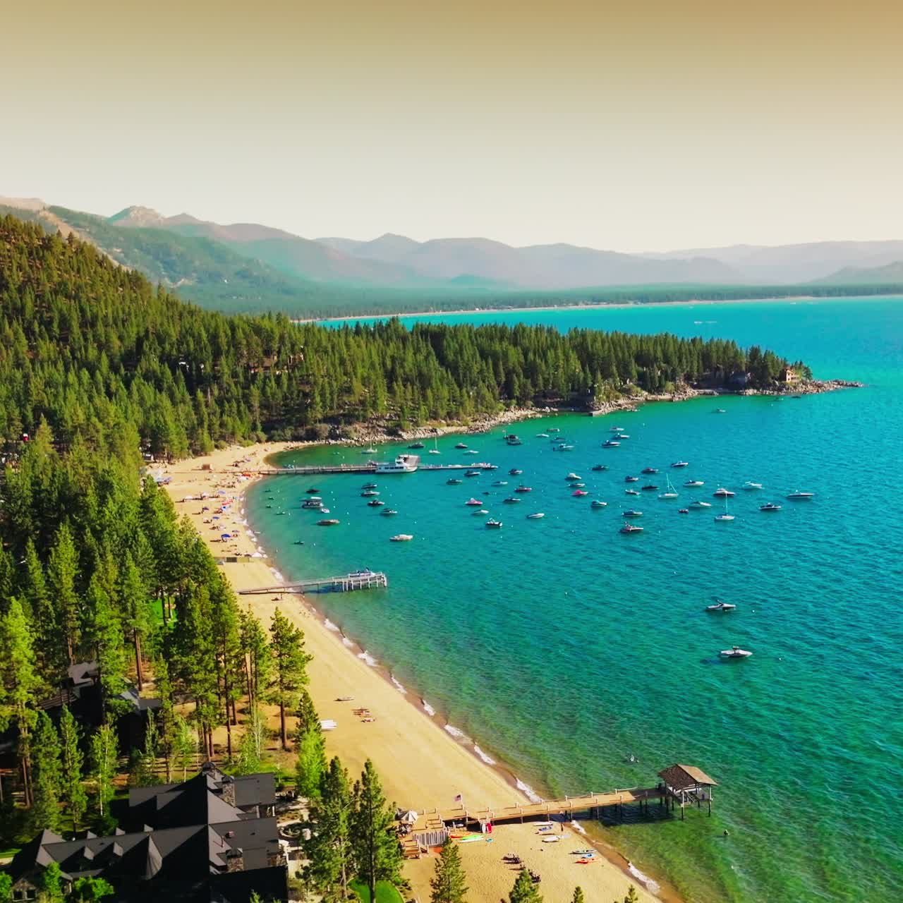 Smuk udsigt over den vidunderlige, blå Lake Tahoe med både ved kysten. Solrig sandstrand og fyrreskov ved vandet. Diset bjerge i baggrunden.
