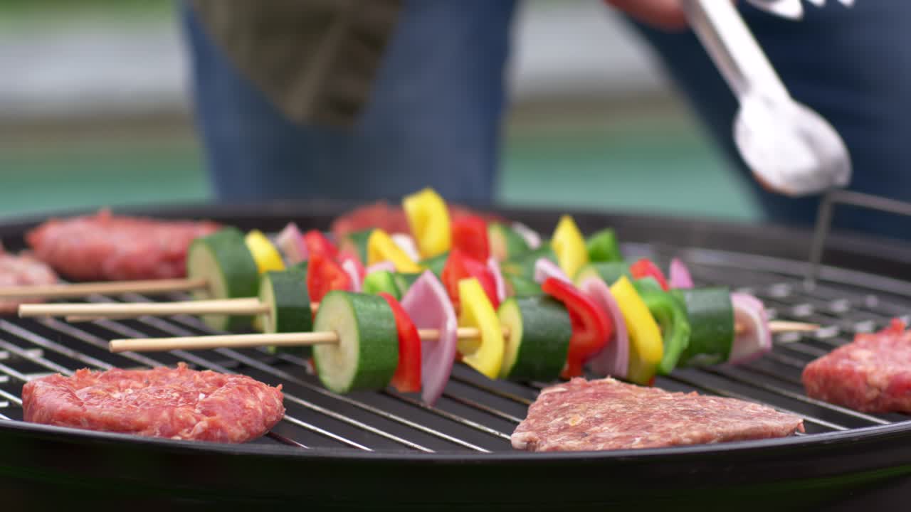 Video verticale: Cuoco con il grembiule che, sollevando le pinze, sposta gli spiedini e preme gli hamburger in giardino per una cottura uniforme