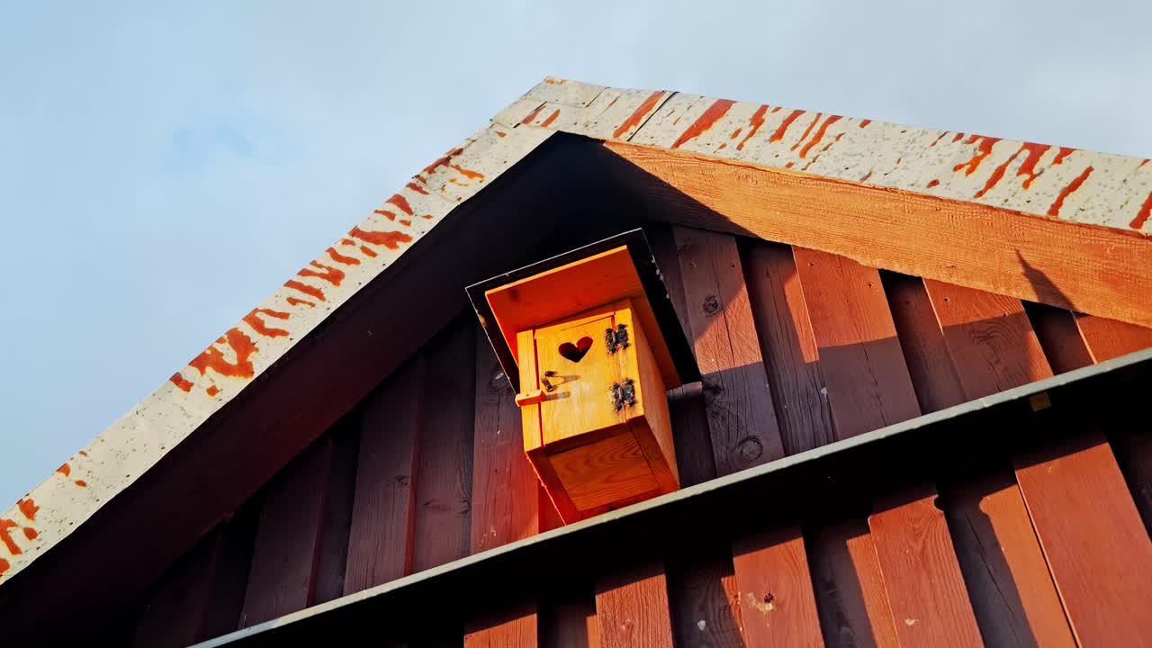 Wooden birdhouse with heart cutout glows in sunlight, ustic gable wall exterior