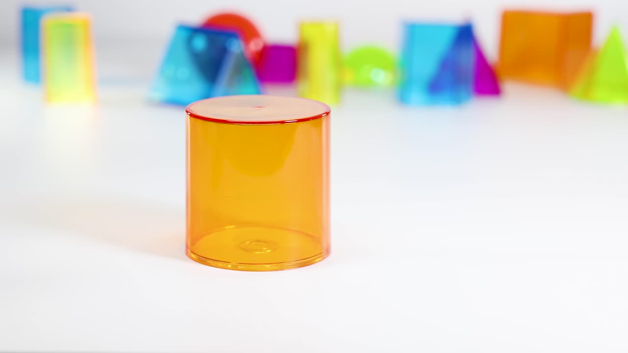 A hand interacts with a translucent orange cylinder among colorful geometric shapes on a white surface