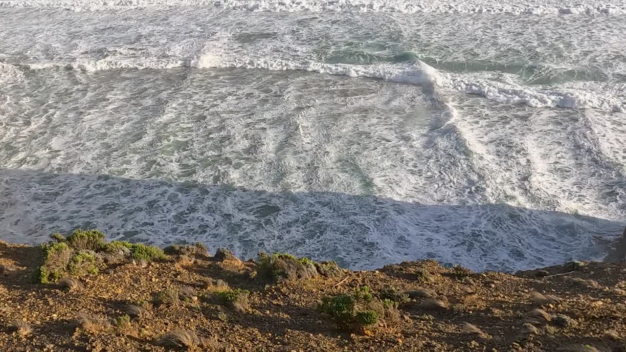 Ocean waves gently roll onto a rocky shoreline, creating a serene coastal scene with sunlight casting shadows.