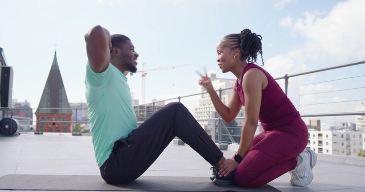 African woman holding anchor and counting reps while African American man doing sit-ups on rooftop