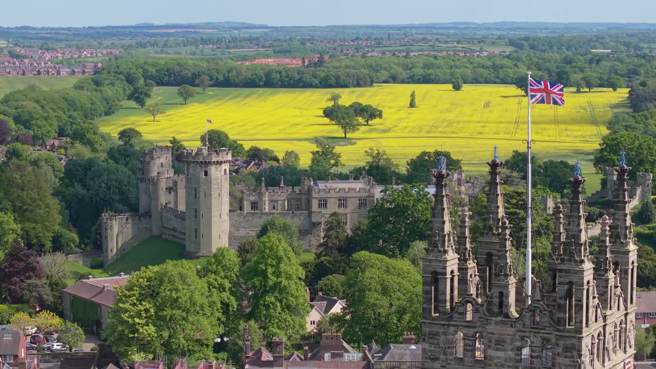 Revelación con dron del Castillo de Warwick, moviéndose desde la Mazmorra hasta el Campanario de Santa María con la bandera de Inglaterra ondeando, mostrando la arquitectura medieval y el paisaje del pueblo circundante.