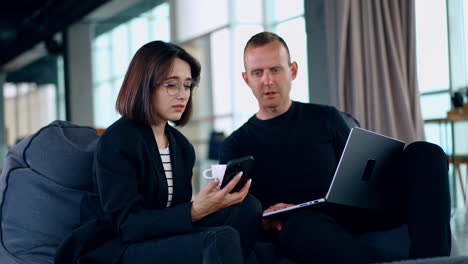 Two colleagues sit comfortably in bean bag chairs talking. Man and woman look at smartphone speaking.