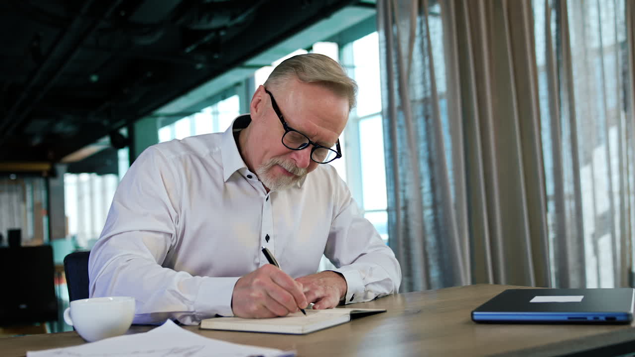 Man in white shirt and glasses sits at desk writing. Man puts his pen into the cup trying to make it write but it doesn't help.