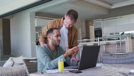 Pareja-Masculina-Gay-Multiétnica-Sentada-A-La-Mesa-En-Casa-Usando-Una-Computadora-Portátil