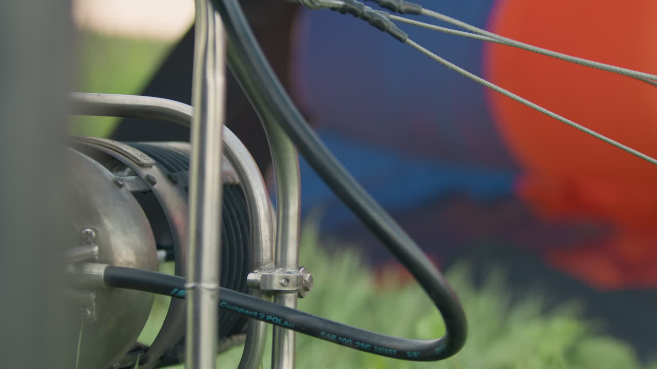 Detailed view of metallic hot air balloon burner mechanism rotating as tall grass gently sways in wind, with colorful deflated balloon blurred in background during open air flight preparation