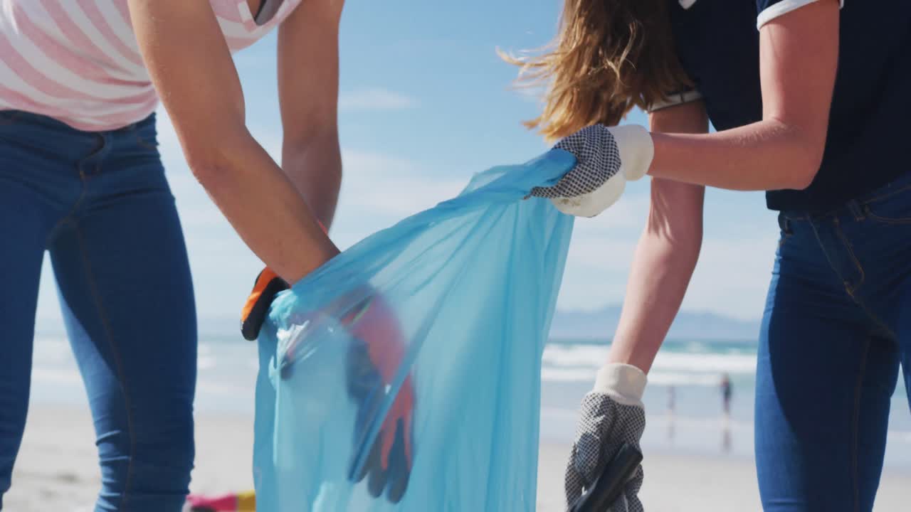 두 명의 다양한 여성 친구들이 해변에서 쓰레기 봉투에 쓰레기를 넣고 있습니다.