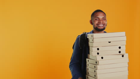 male pizzeria courier carrying huge pile of pizza boxes in studio