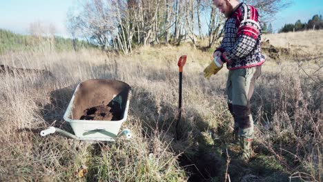 a man excavates soil and transfers it into a wheelbarrow for the construction of a greenhouse - static shot