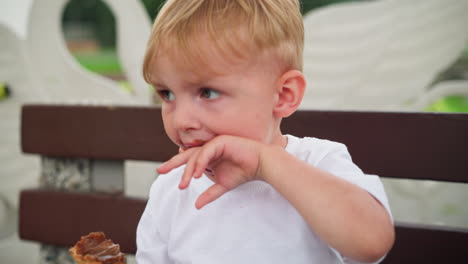 un niño pequeño, sentado en un banco, tiene chocolate manchado alrededor de su boca mientras parece ligeramente confundido