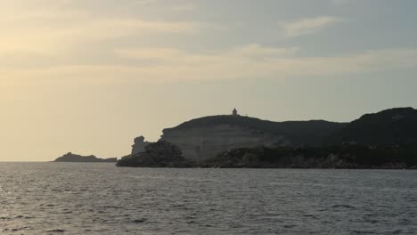 punto de vista de la proa del velero navegando sobre el agua de mar ondulada hacia los acantilados de la isla de córcega y el faro de capo pertusato en francia, cámara lenta