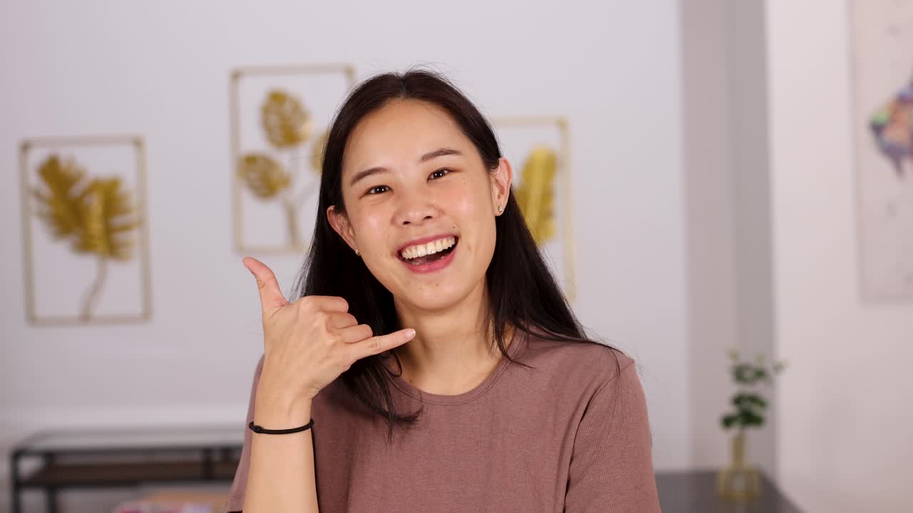 Asian woman smiles and makes a call sign gesture with her hand in a brightly lit, modern indoor setting with soft, natural lighting