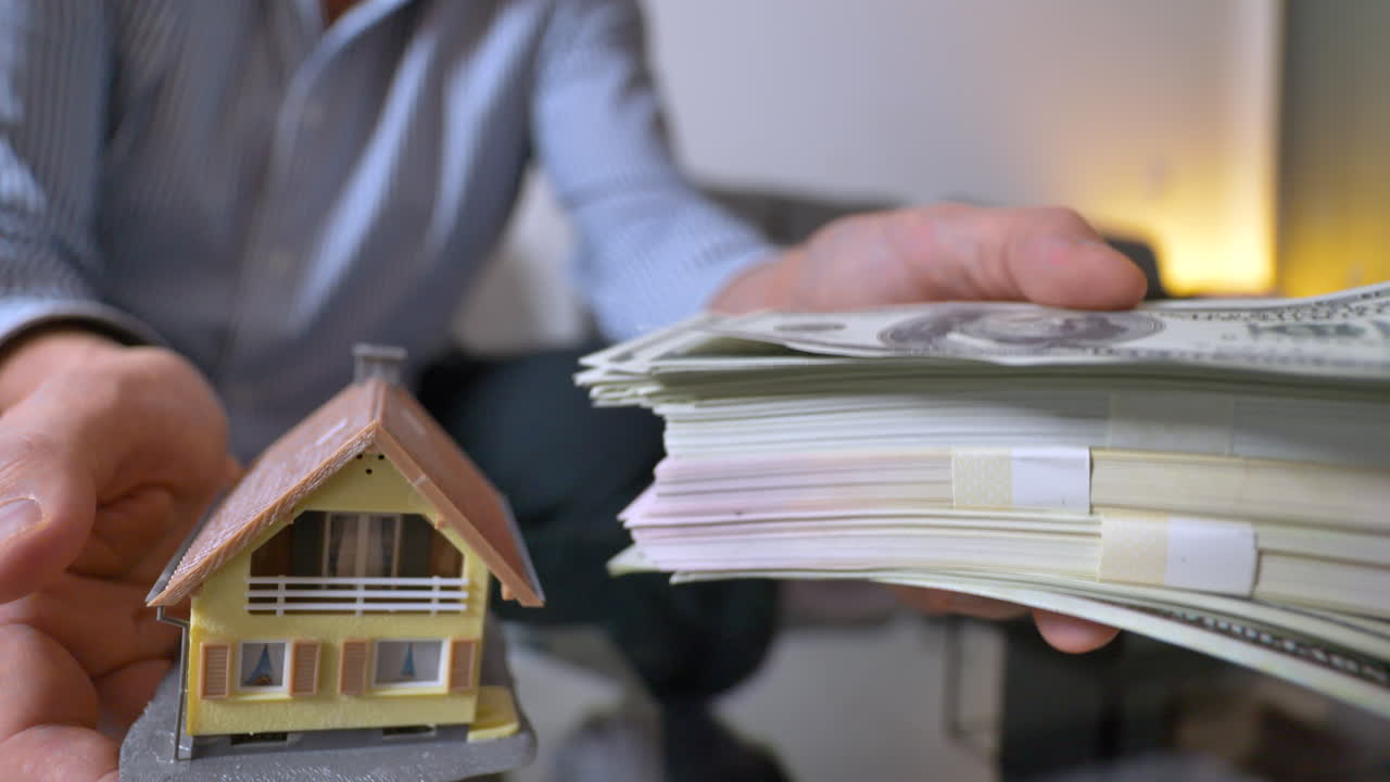 Primer plano de un hombre adulto levantando una casa o un fajo de billetes en casa: consideración de dinero en efectivo o inversión inmobiliaria.