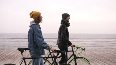 Side-View-Of-Smiling-Couple-Of-Young-Hipsters-Walking-Together-With-Their-Bikes-Near-The-Sea-At-Autumn-Day