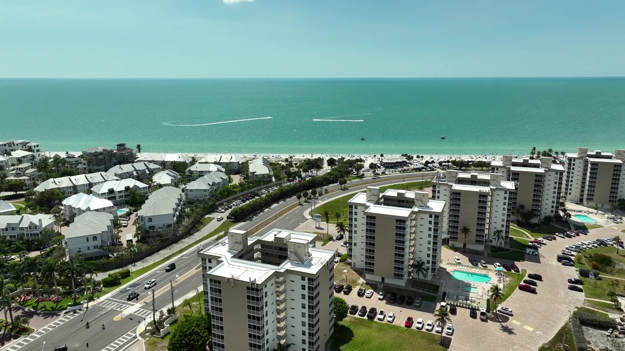 Vista aérea cinematográfica de Bonita Beach Road en Bonita Springs, Florida, mostrando la concurrida calzada, lujosas mansiones frente al mar, canales de manglares y propiedades junto al Golfo.
