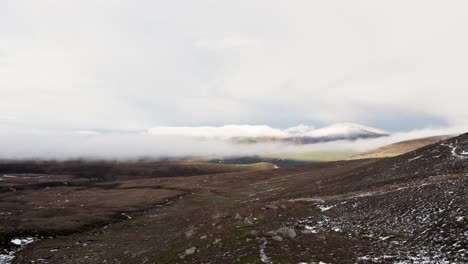 Aerial-drone-footage-in-winter-in-Coire-an-t-Sneachda-in-the-Cairngroms-National-Park-in-Scotland-with-patchy-snow,-mountains-and-clouds-moving-forwards-towards-Aviemore-and-Glen-More-with-an-icy-path