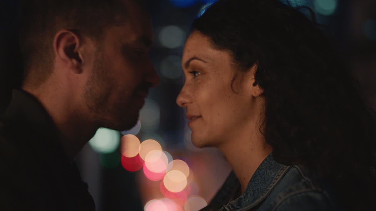 젊은 백인 커플이 밤에 지붕에서 키스하고 평화로운 도시 저녁에  ⁇ 만적인 관계를 즐기고 있습니다.