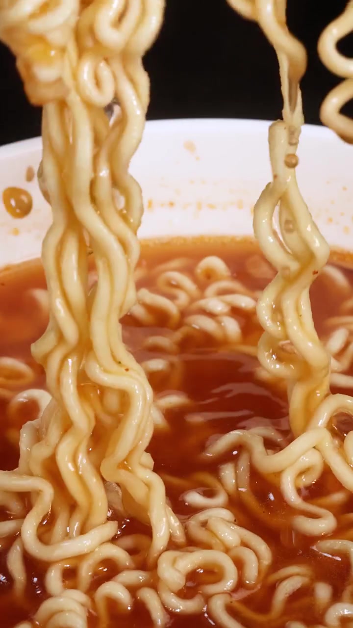 Noodles istantanei cotti vengono rilasciati dalle bacchette in una ciotola di brodo rosso, catturati in primo piano con illuminazione brillante e uniforme e movimento minimo della fotocamera