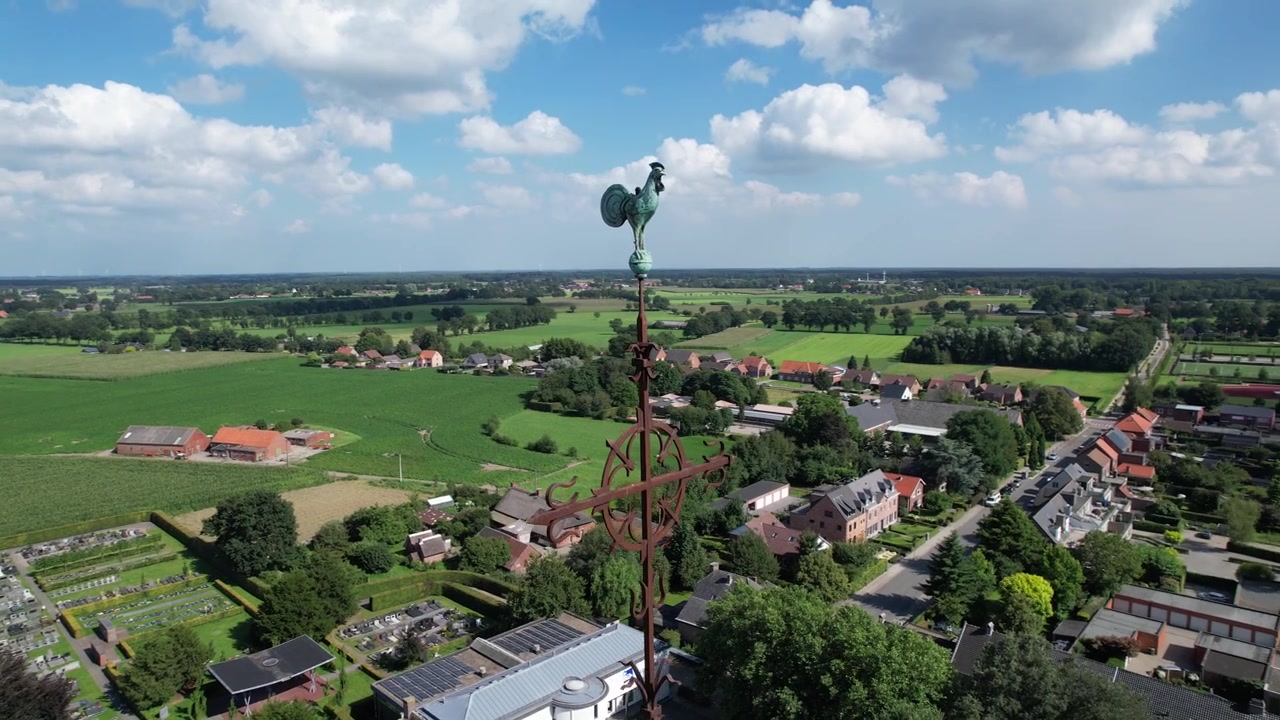 Bir kilise kulesi horozundan yükselen drone görüntüsü, dağınık bulutlu parlak bir gökyüzü altında yeşil tarlalarla çevrili kırsal bir köyü ortaya çıkarıyor