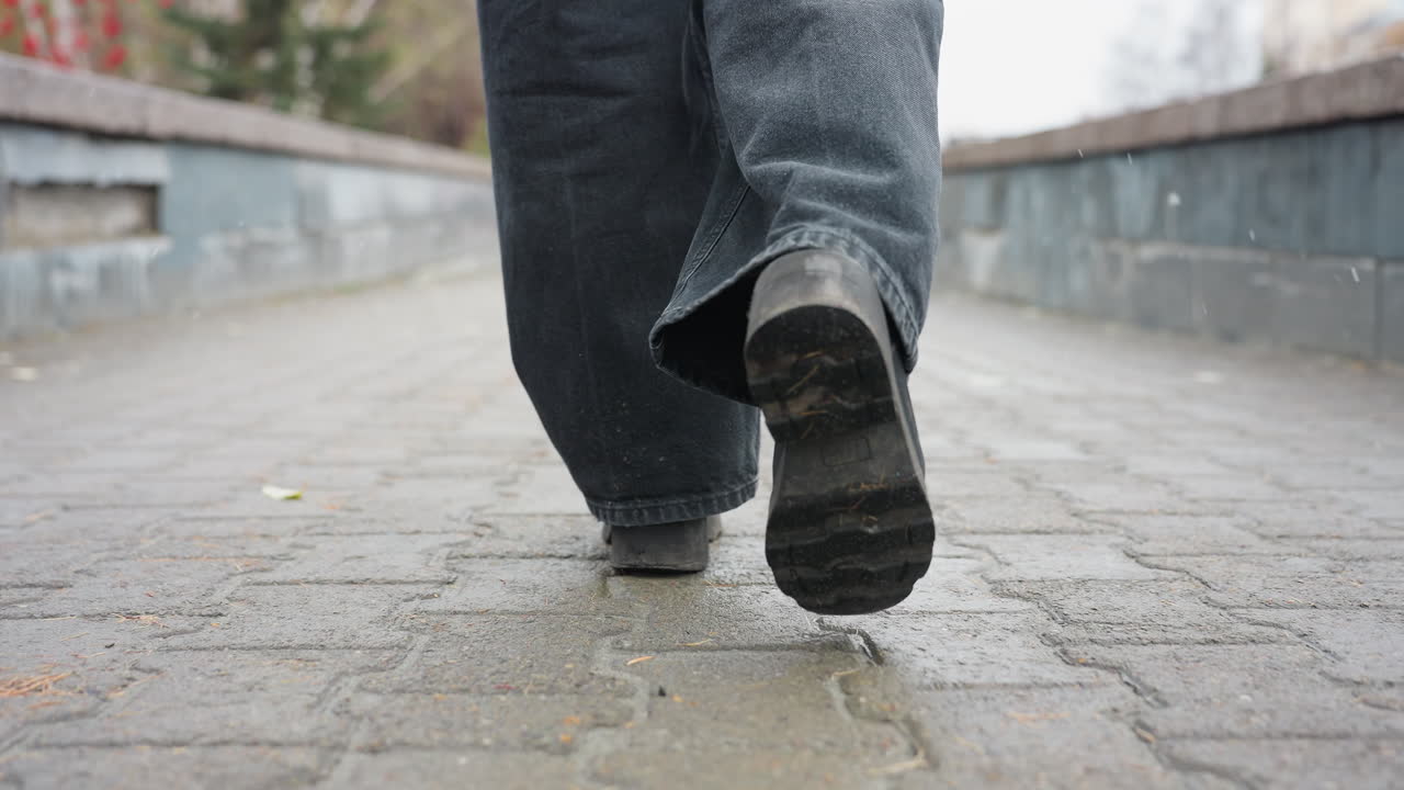 Back shot of person's legs in black trousers and black boots walking slowly on outdoor paved path during calm overcast day, capturing subtle foot movement and rhythm in peaceful urban setting