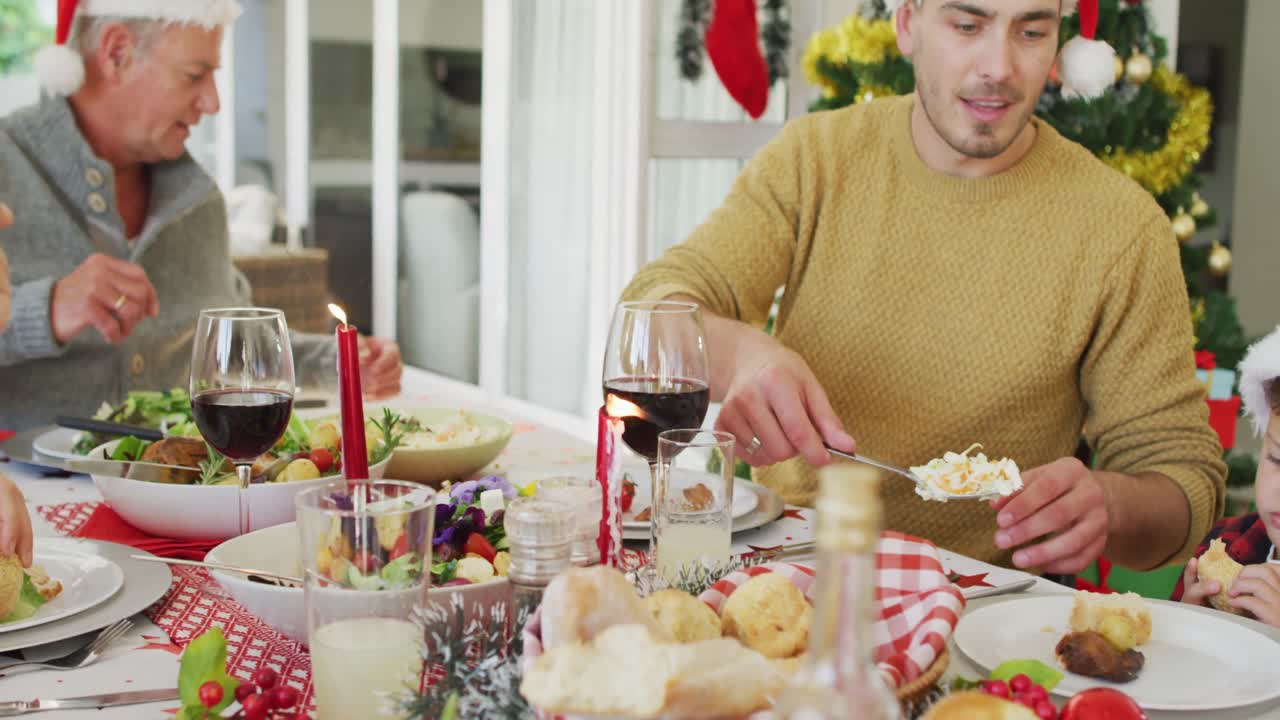 familia caucásica feliz de varias generaciones con sombreros de papá noel, teniendo la comida de navidad