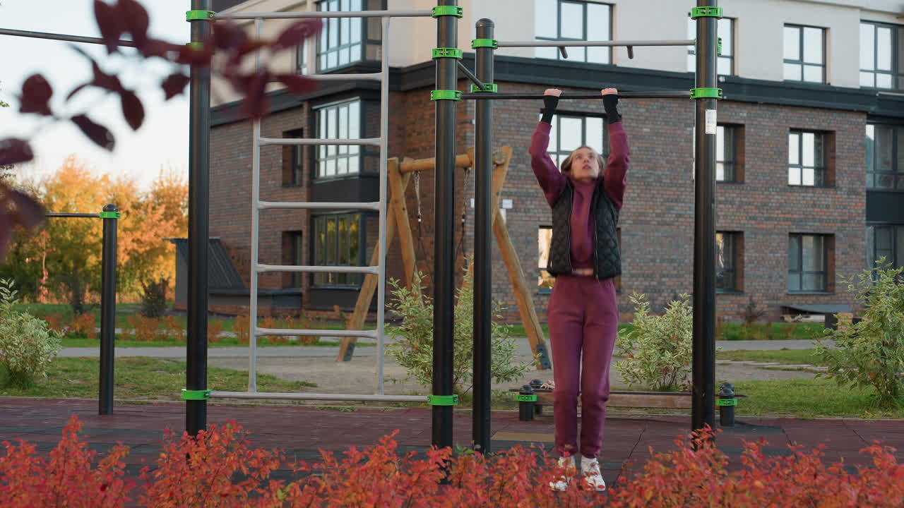 Fitness trainer working out on horizontal pull up bar in park setting with autumn foliage backdrop and city building background, demonstrating forearm strength, and intense focus