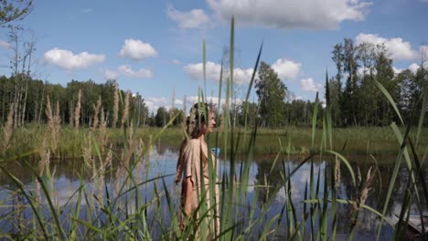 a woman is walking by the pond as the camera is following her with tall grasses in the foreground, natural colour, slow motion