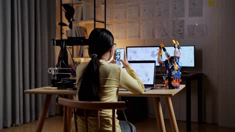 back view of teen asian girl with 3d printing looking at the pictures on smartphone while designing a cyborg hand on a laptop at home