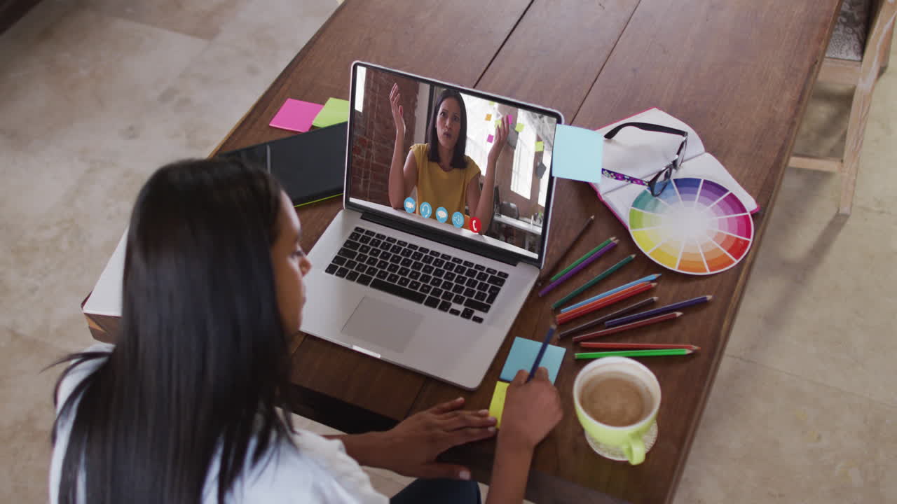 Empresaria birracial sentada en una mesa usando una computadora portátil teniendo una videollamada con una colega.