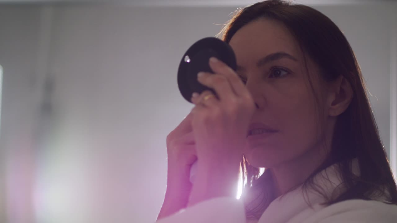 작은 주머니 거울을 들고 집 화장실에서 화장을 하는 갈색 머리 여자