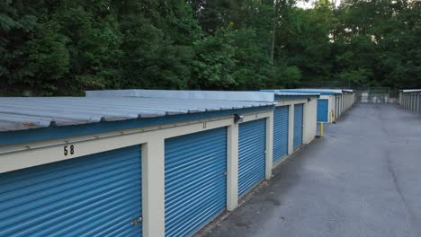 A-row-of-blue-storage-units-with-white-trim,-numbered-doors,-and-a-paved-pathway,-surrounded-by-trees