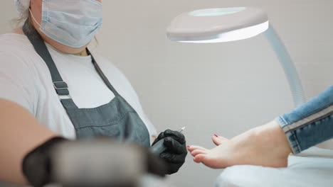 close up of nail technician wearing mask and gloves connecting electric filer tool while client rests foot with red polish on padded surface under bright lamp in clean professional salon environment