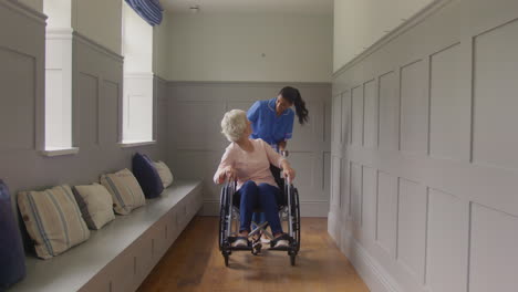 Senior-Woman-At-Home-Being-Pushed-In-Wheelchair-By-Female-Care-Worker-In-Uniform