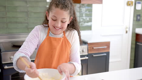 Happy-biracial-girl-with-long,-curly-hair-mixing-dough-in-bowl-and-smiling-in-sunny-kitchen