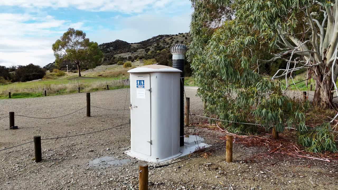Drone camera smoothly pans around a small standalone public toilet in a gravel clearing, surrounded by trees and natural landscape under daylight