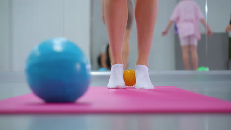 lower leg view during a workout session with a small yellow ball between ankles and a blue ball in front, large mirror reflecting other participants in the background