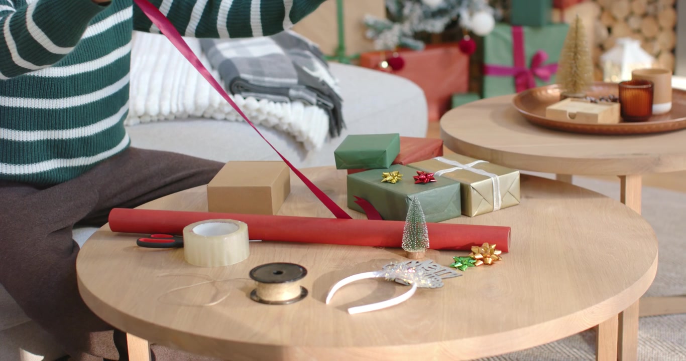Person is reaching, pulling ribbon from spool to drape over box while wrapping gifts on table