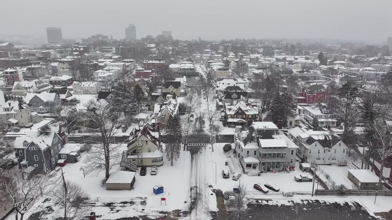 Triple-deckers line a quiet road in Worcester, Massachusetts. Downtown towers rise over neighborhood rooftops. Sweeping above city blocks, aerial.