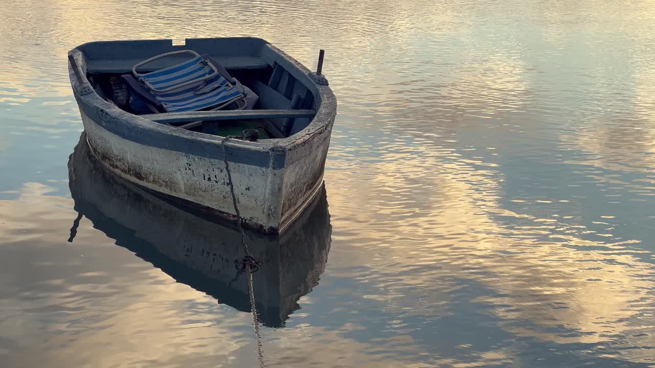 маленькая рыбацкая лодка с сложенным пляжным стулом внутри, мягко плывущая по спокойной воде барбате, андалусия, отражая теплые оттенки неба во время мирного заката