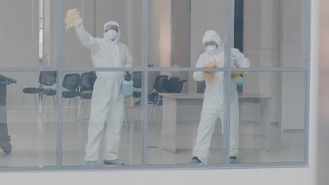 view from outside of two cleaning men wearing personal protective equipment cleaning window panes inside an office building