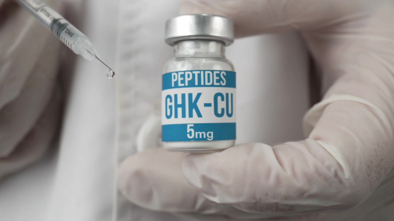 Mani guantate tengono una fiala di peptide GHK-CU mentre una siringa si avvicina con una gocciolina visibile, suggerendo iniezione o uso per ricerca clinica