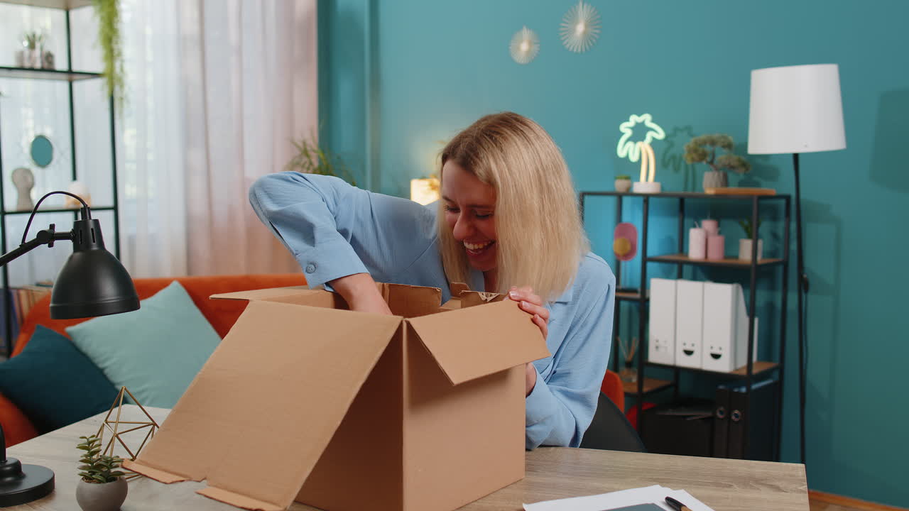 Happy blond businesswoman unpacking opening delivery parcel box purchase sits at desk in apartment