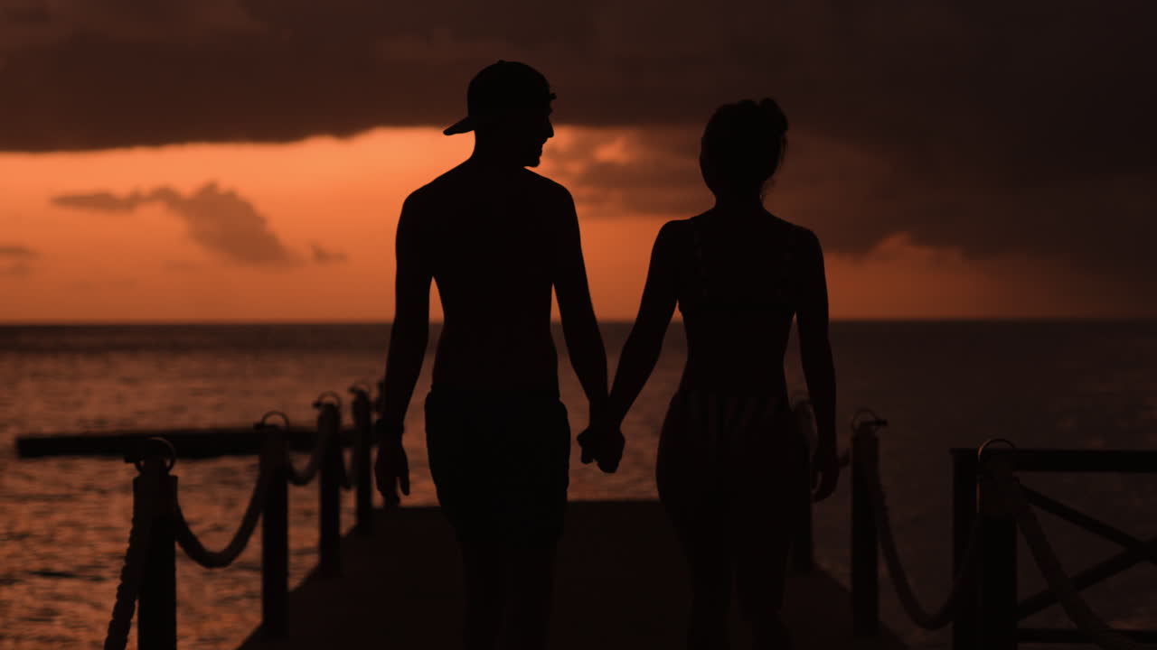 Couple walks hand in hand at sunset, Romantic sunset walk on a dock, couple silhouette against a background of calm sea horizon, with orange fiery clouds